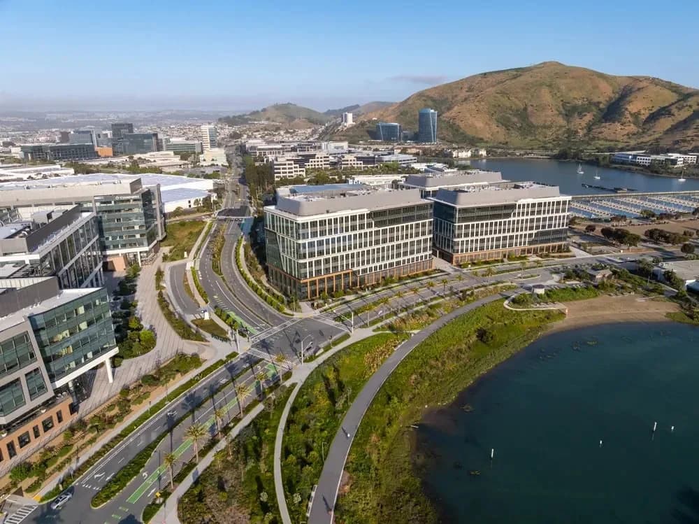 Aerial view of a modern waterfront office campus with a curved road and hills in the background