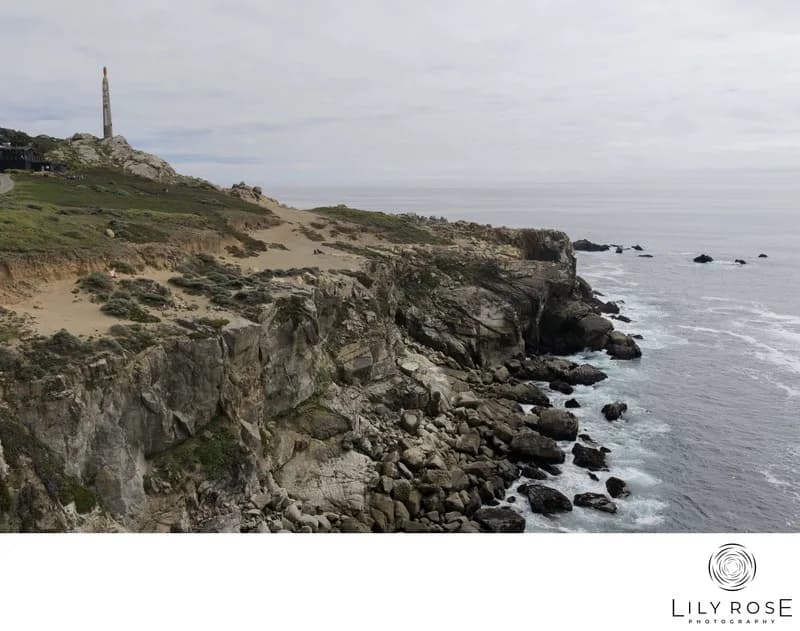 Rocky coastline with cliffs and a tall hilltop tower, waves along the shore, Lily Rose Photography logo right