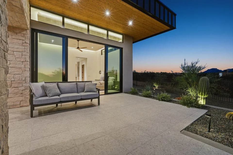 Patio with outdoor sofa and sliding glass doors of a modern home at dusk, desert landscaping and illuminated cactus