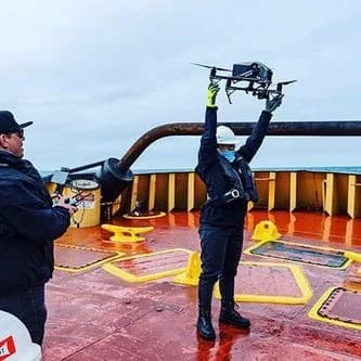 Two crew members on a red ship deck; one lifts a drone overhead, other holds controller; sea in background