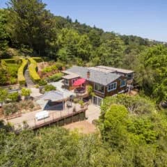 Drone view of a two-story house with a deck, red umbrella, and surrounding trees
