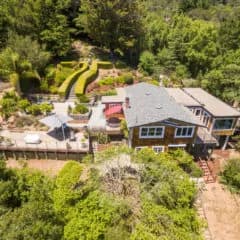 Aerial view of a two-story house with a curved lime-green lawn, red umbrella on a deck, and trees