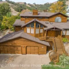 Aerial view of a large brown wooden house with multiple rooflines, attached garage, deck, and hillside trees