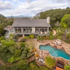 Two-story house with wraparound balcony, surrounding trees, and a curved pool with stone patio