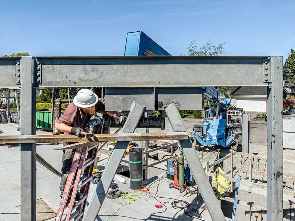 Construction worker with white helmet and gloves on a ladder amid steel framing and machinery outdoors