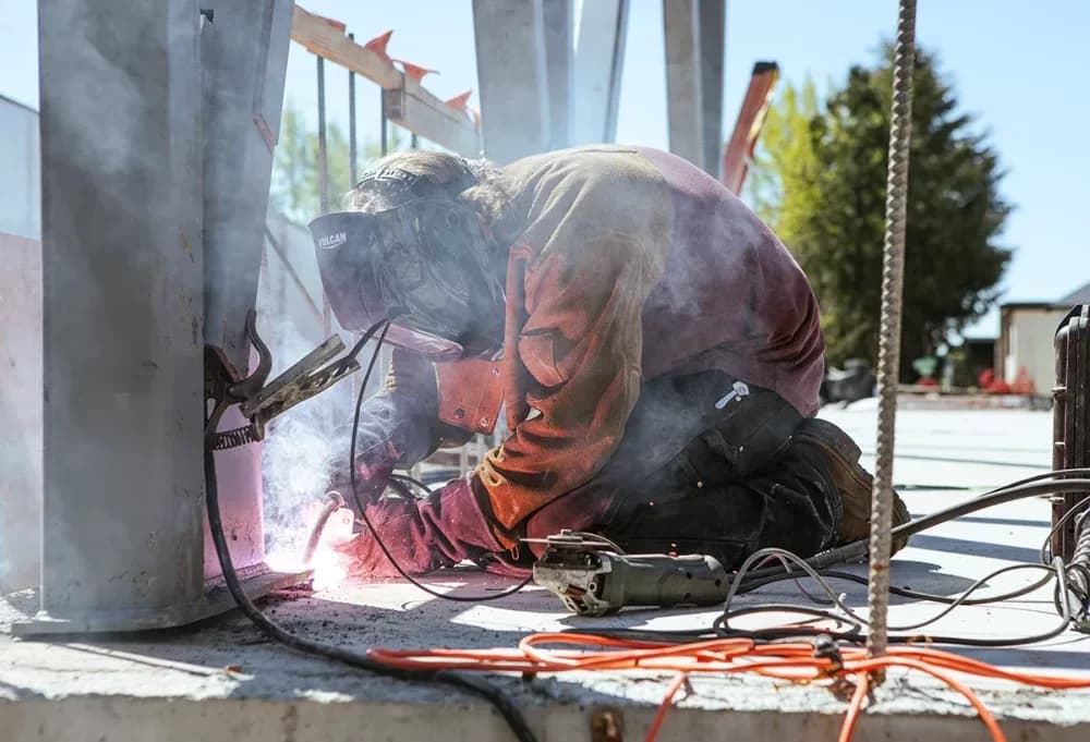 Welder wearing a welding helmet kneels on a concrete surface, welding a metal beam with sparks