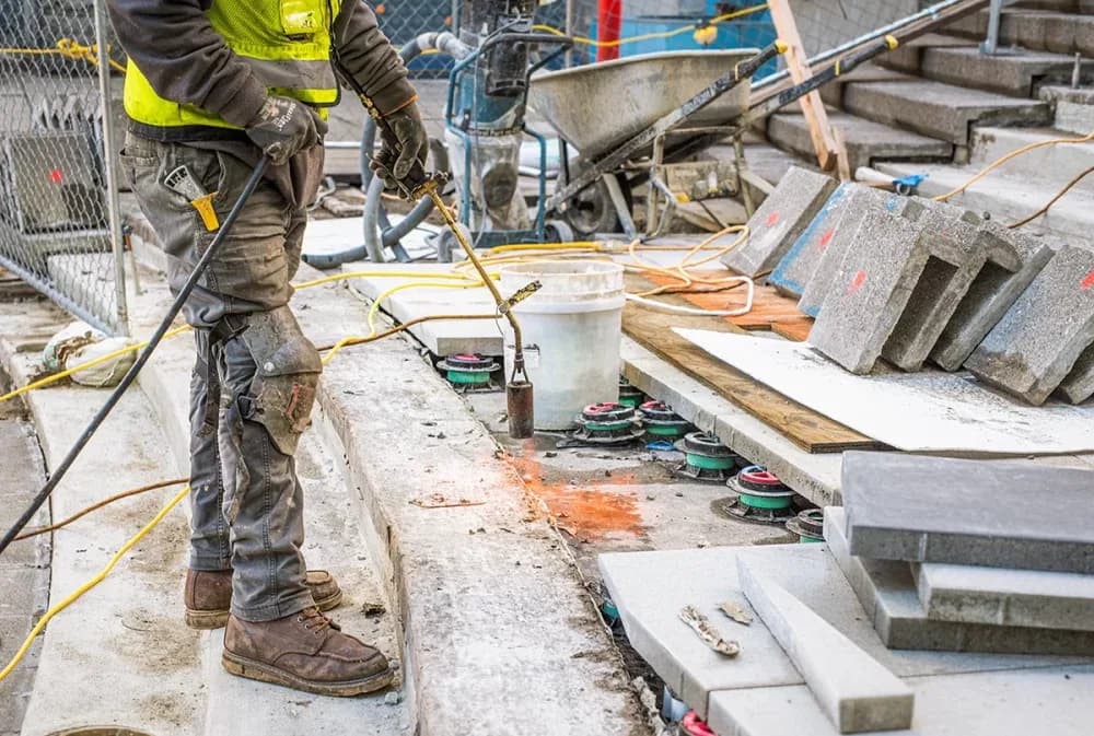 Construction worker in a yellow safety vest uses a tool by a curved concrete edge with blocks and buckets