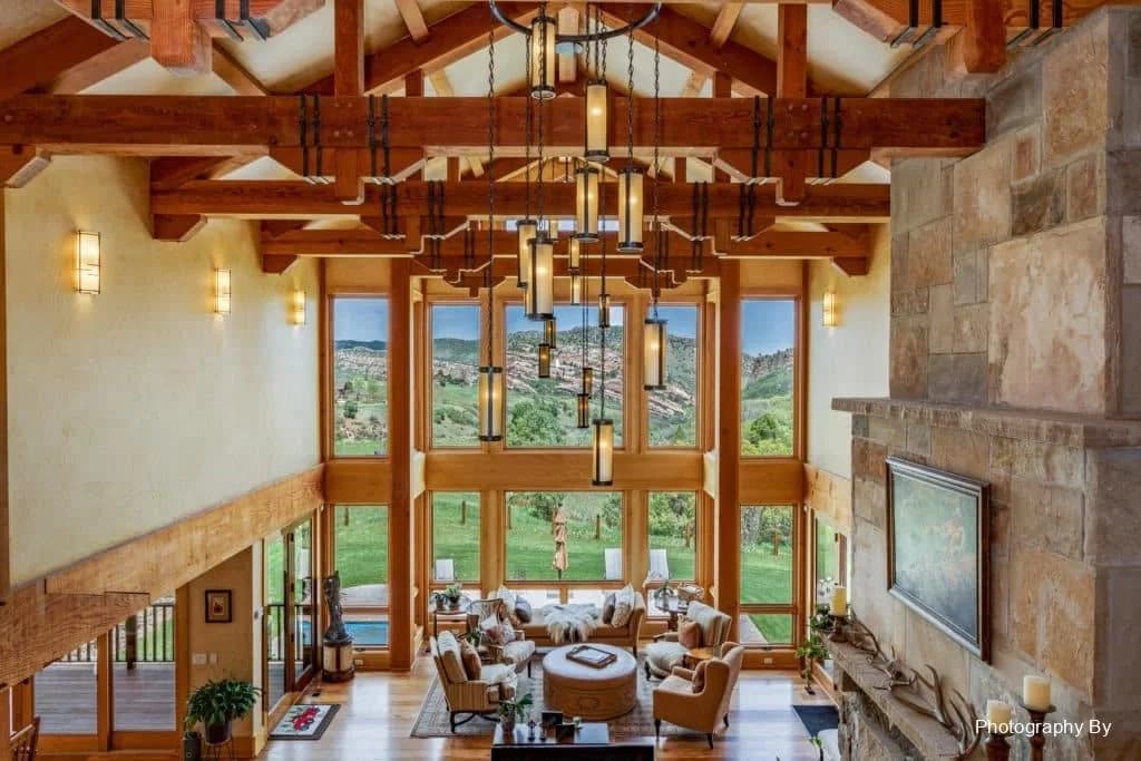 Living room with exposed wooden beams, chandeliers, stone fireplace, and large windows with a countryside view