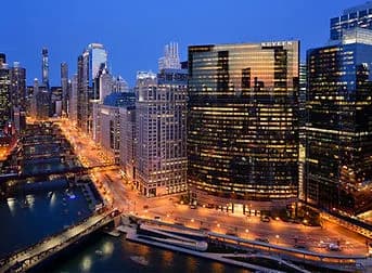 City skyline at dusk with illuminated high-rise buildings along a river, a bridge and boats
