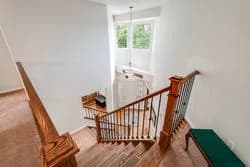 Interior view of a two-story stairwell with wooden banisters, a white cabinet by a window, and a chair
