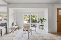 Bright white living area with a wooden dining table, chairs, gray sofa, rug, and a potted plant by windows