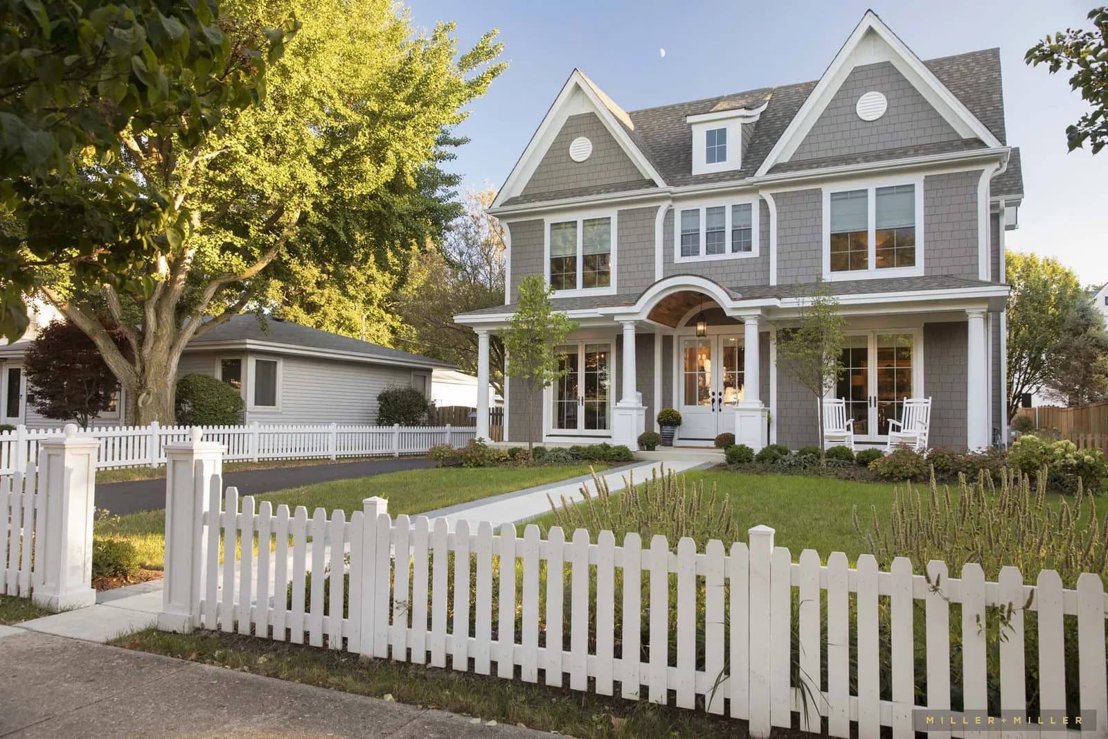 Two-story gray house with white trim, front porch with arches and white columns, and a white picket fence