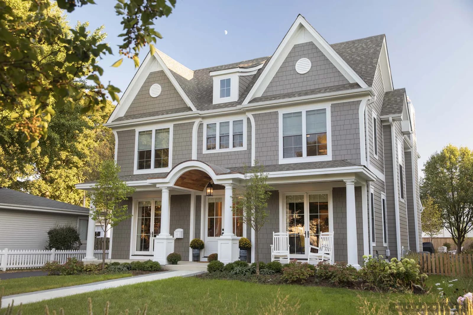 Two-story gray house with white trim, front porch with arches and columns, rocking chairs