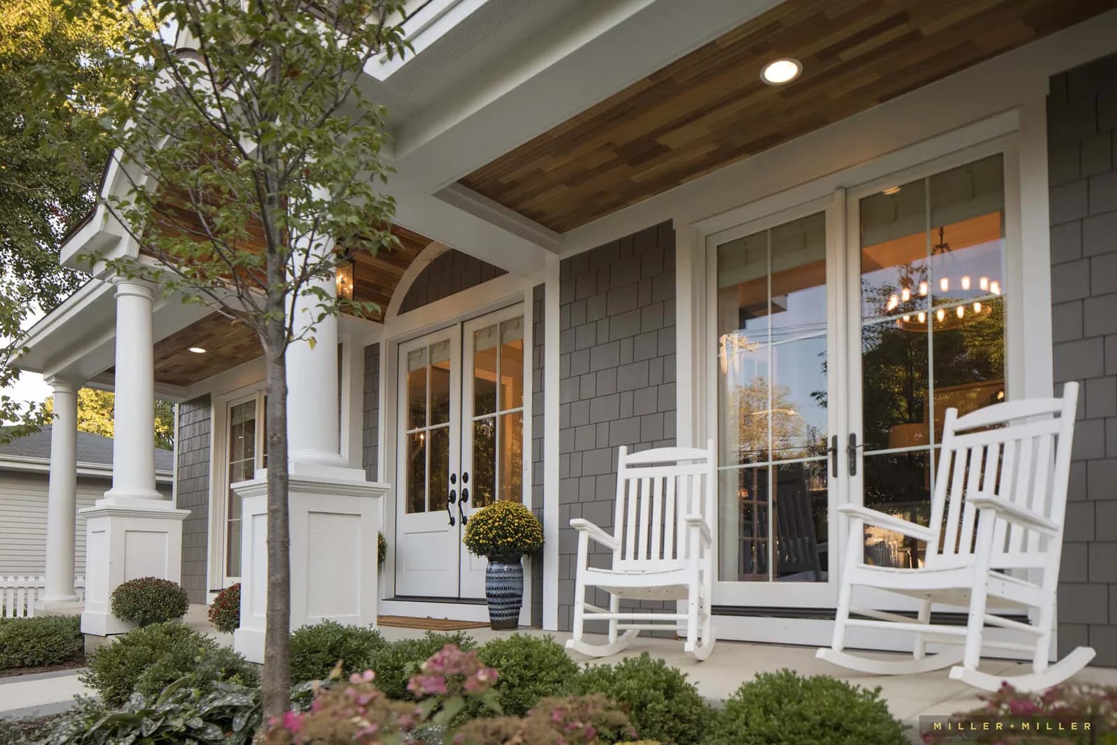 Front porch of a gray shingle house with white columns, glass doors, vase with mums, and white rocking chairs