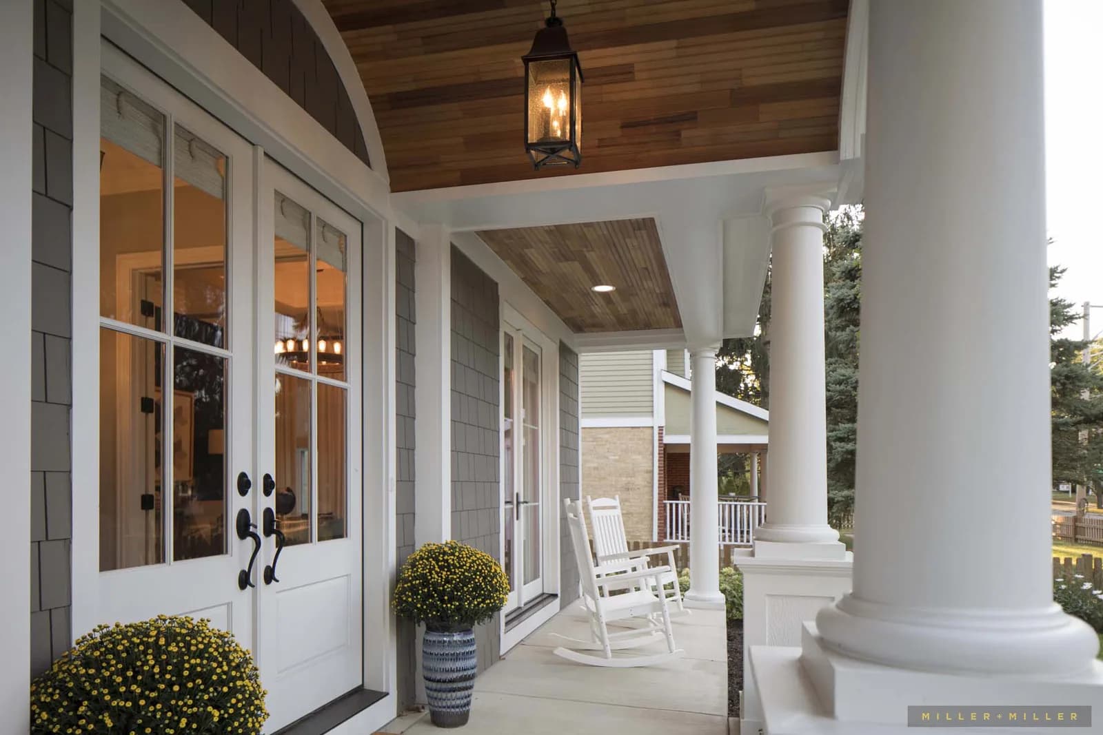 Front porch with white columns, glass panel doors, rocking chairs, lantern light, and potted mums