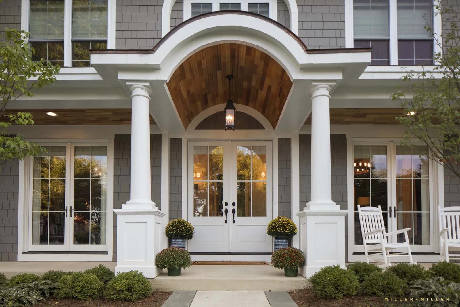 Front porch of a gray shingle house with white columns, arched wood ceiling, glass doors, lantern and plants