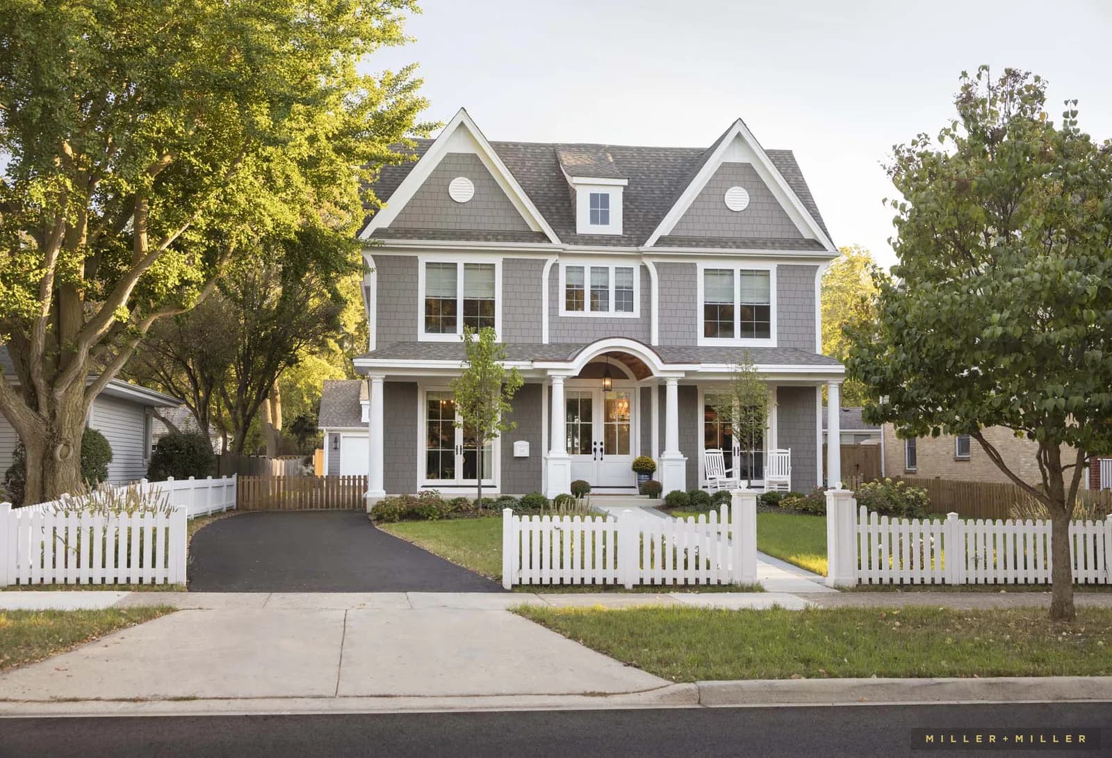 Front view of a gray two-story house with white trim, arched entry, white picket fence, and surrounding trees