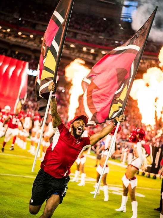 Man running on football field waving two large team flags with cheerleaders and flames behind