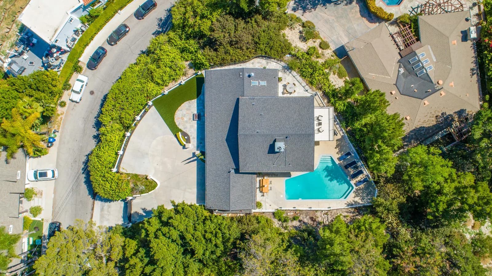 Aerial view of a large house, turquoise pool, curved driveway, and surrounding trees
