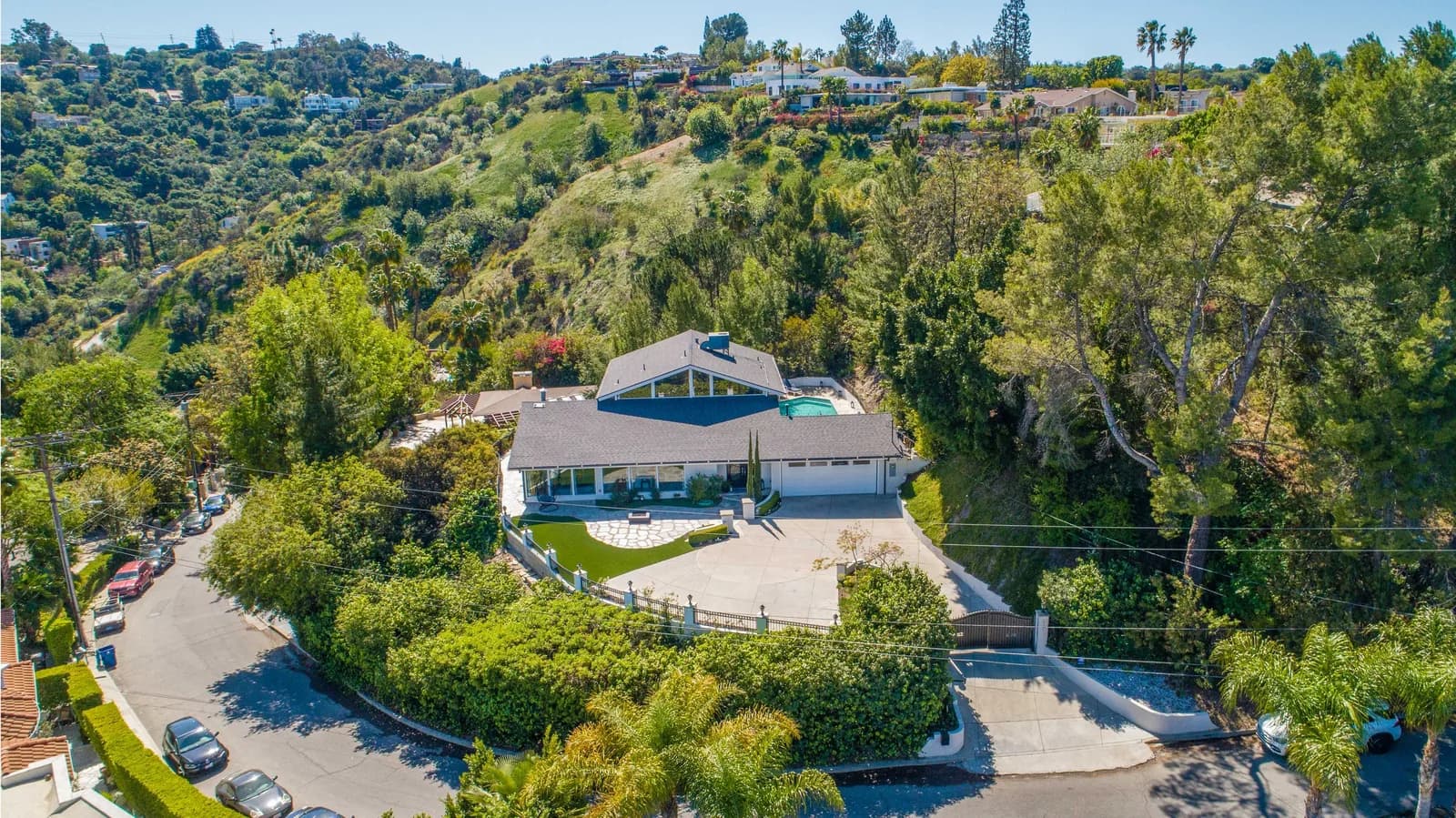 Aerial view of a hillside home with a pool, curved driveway, and surrounding trees