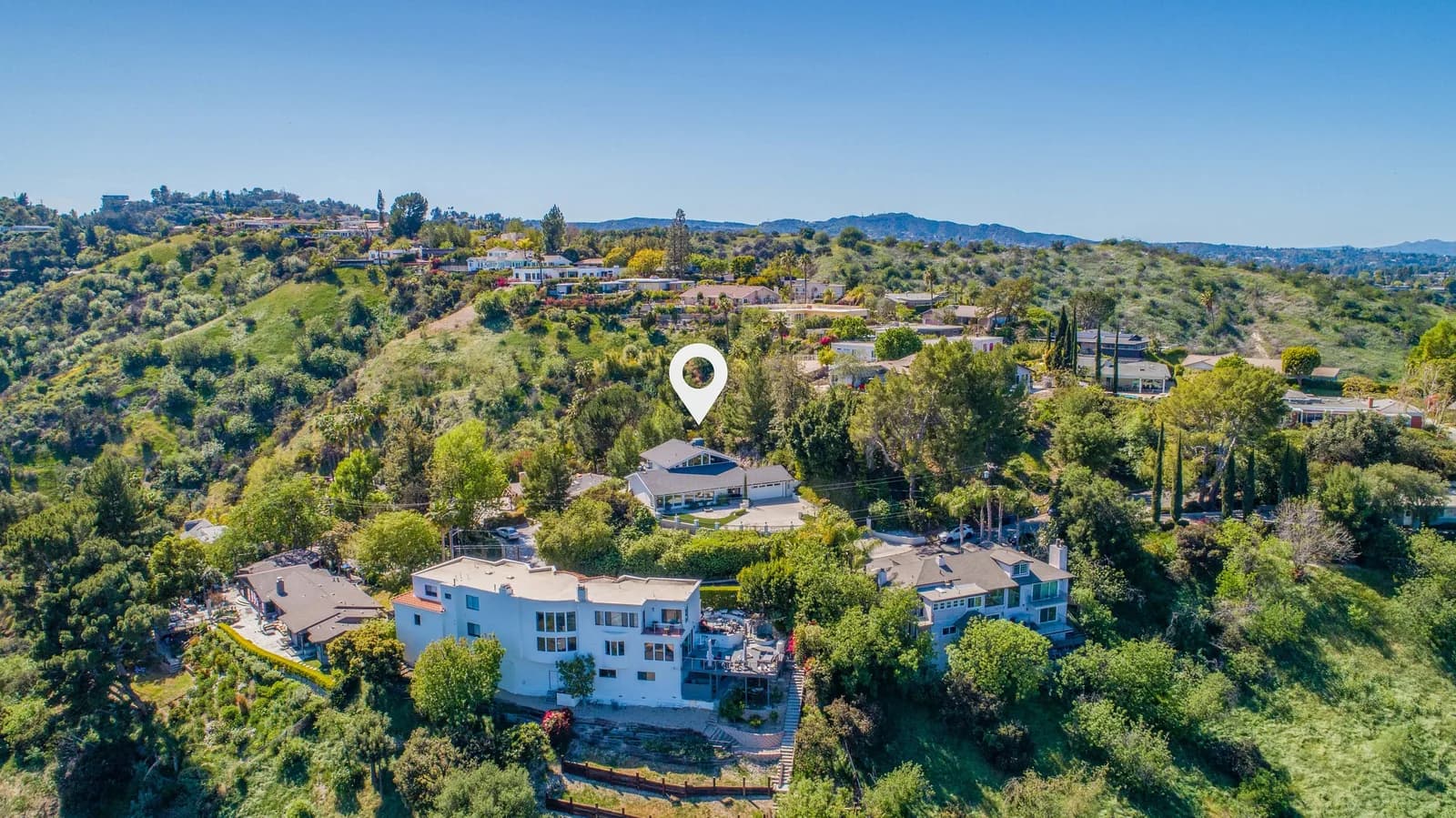 Aerial view of a hillside residential area with trees and houses; a location pin marks a central home