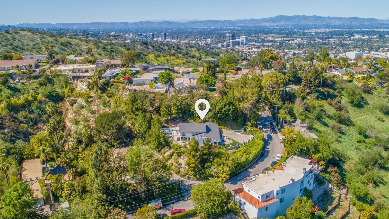 Drone aerial view of a hillside neighborhood with a white location pin above a house and distant cityscape