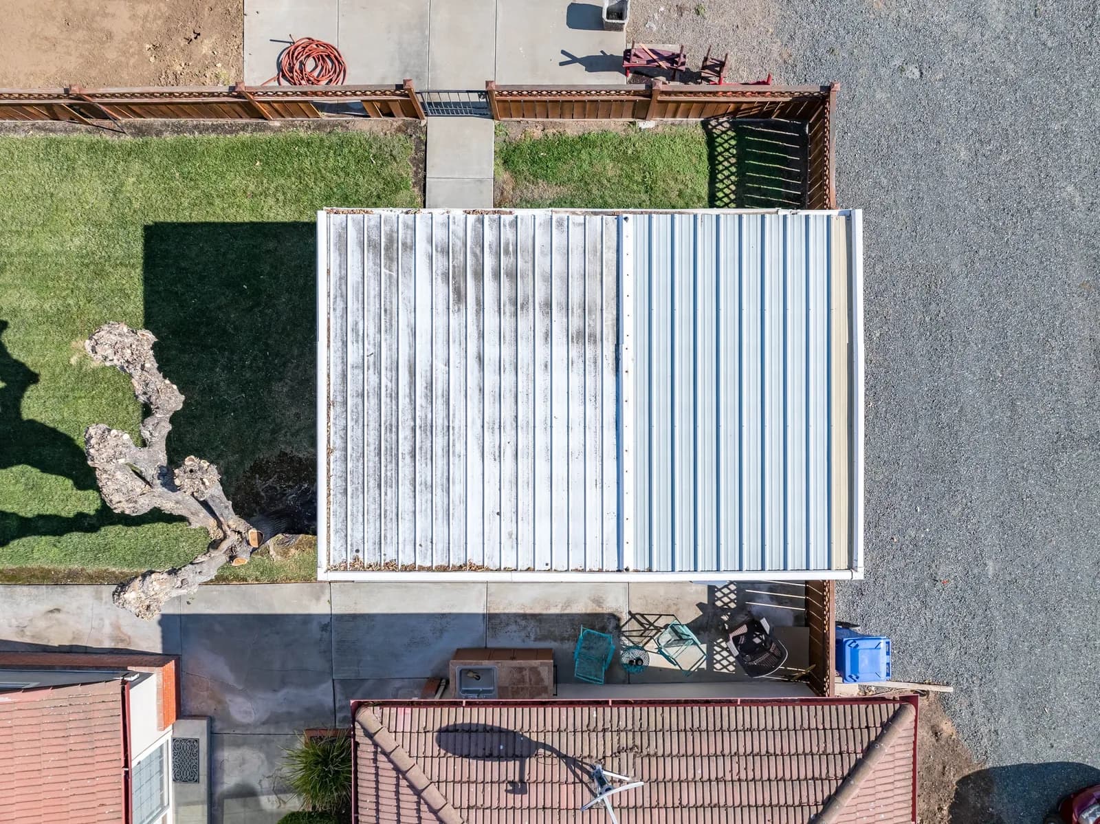 Aerial view of a two-tone corrugated metal roof with a grassy yard, concrete path, wooden fence, and gravel