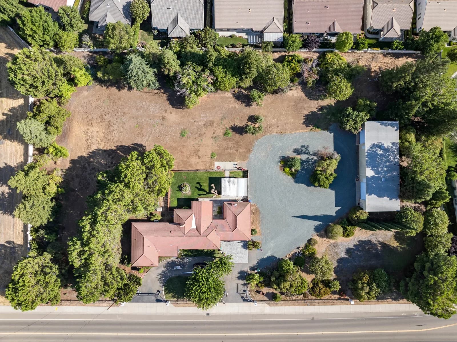 Aerial view of a residential property with red-roof house, gravel area, dirt yard, and surrounding trees