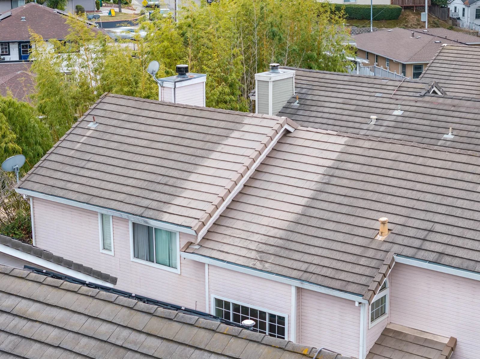 View from above of tan tiled roof lines, two white chimneys, a satellite dish, and nearby houses and trees
