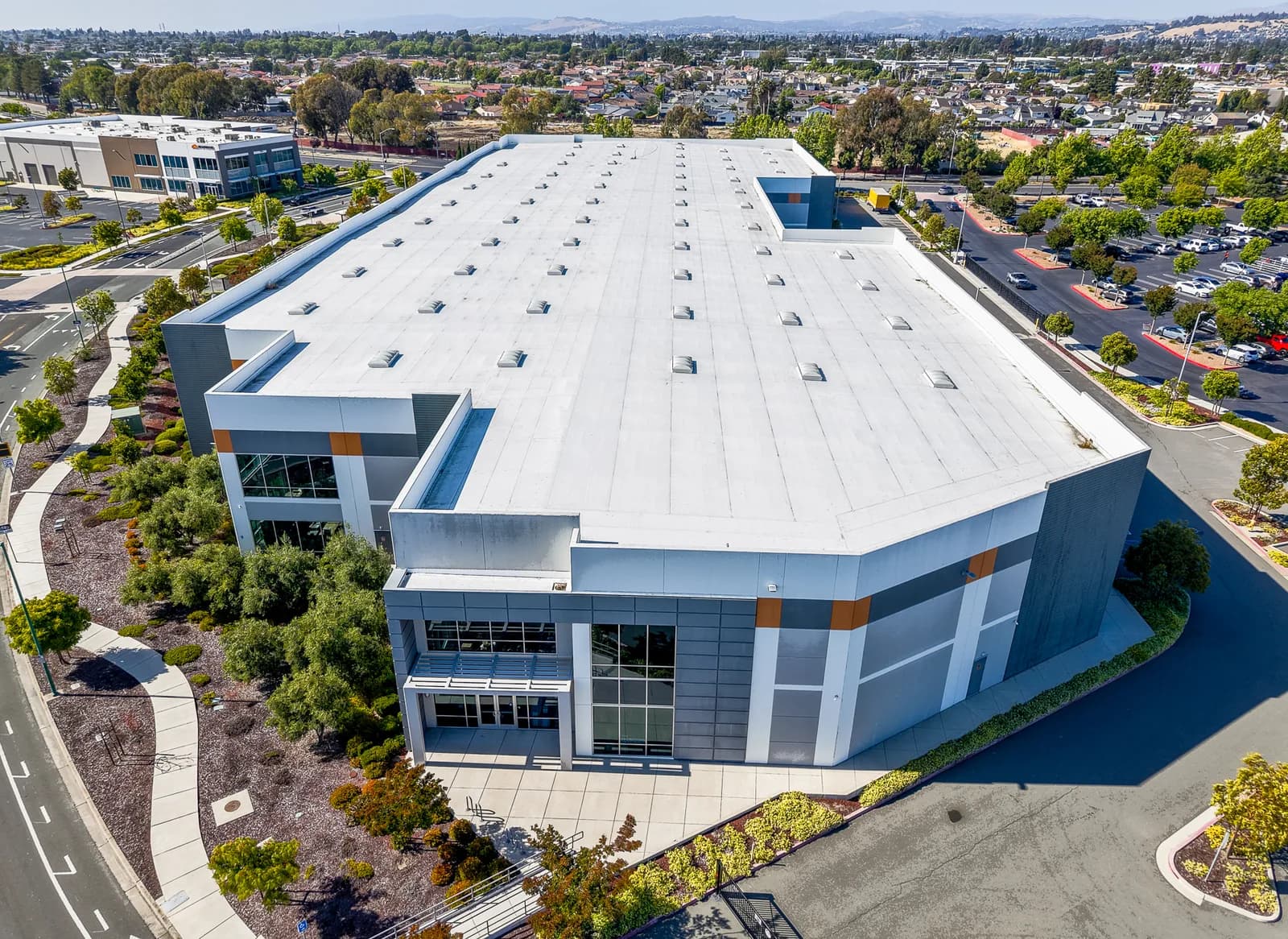 Aerial view of a large modern building with a white flat roof and skylights, surrounded by trees and parking