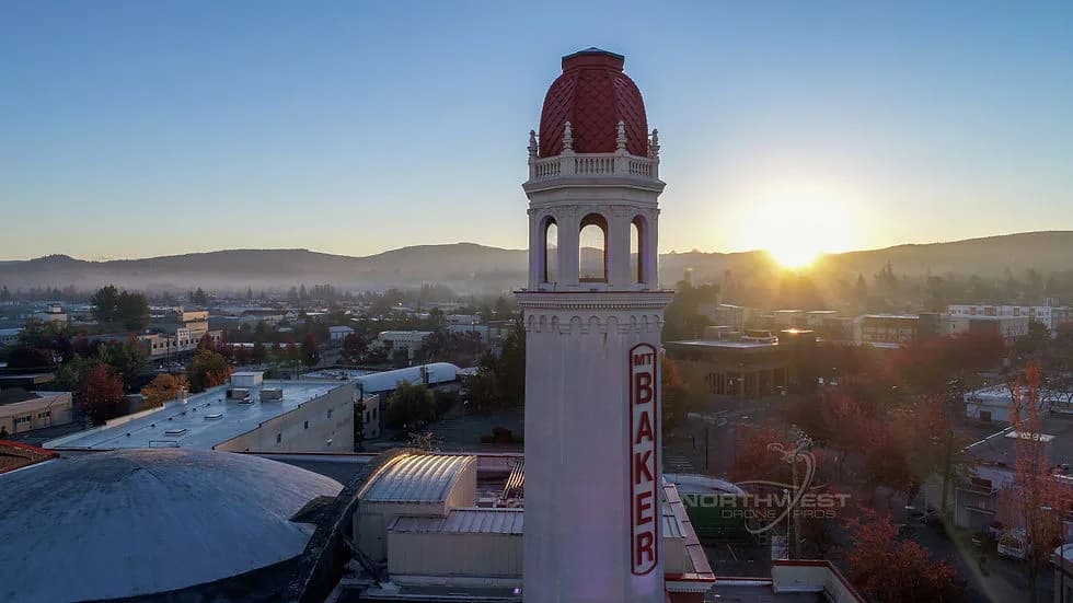 Tall white tower with a red domed top and a vertical sign, in a town at sunset with hills on the horizon