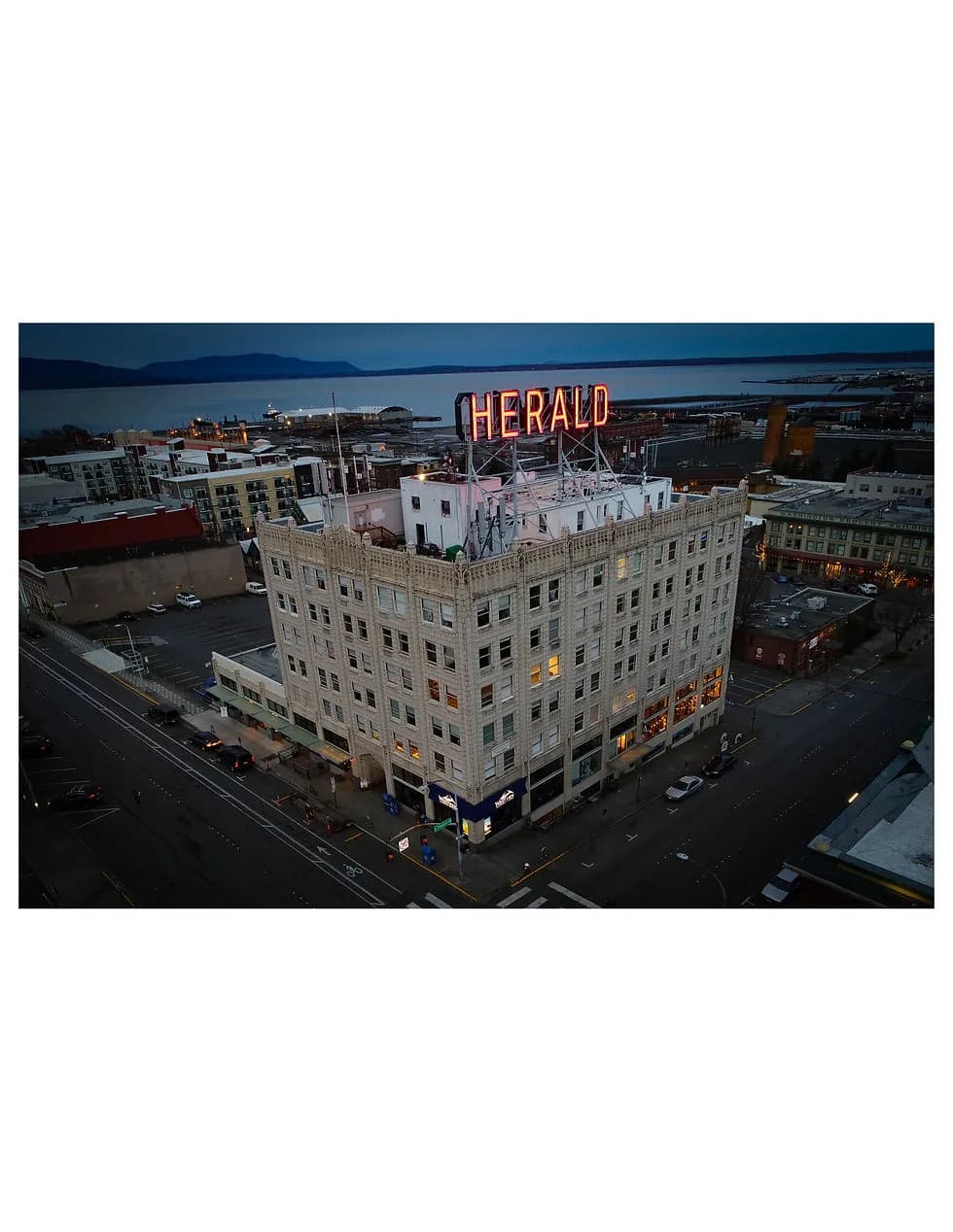 Aerial view of a tall brick building with a neon 'Herald' sign on the roof, downtown streets below
