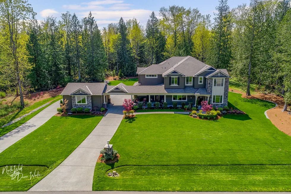 Large two-story gray house with front lawn, curved driveway, and surrounding trees in a suburban setting