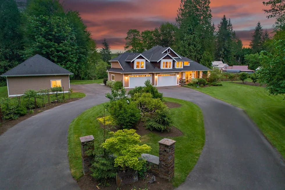A large house with curved driveway, garages, and a separate outbuilding to the left on a green lawn at sunset