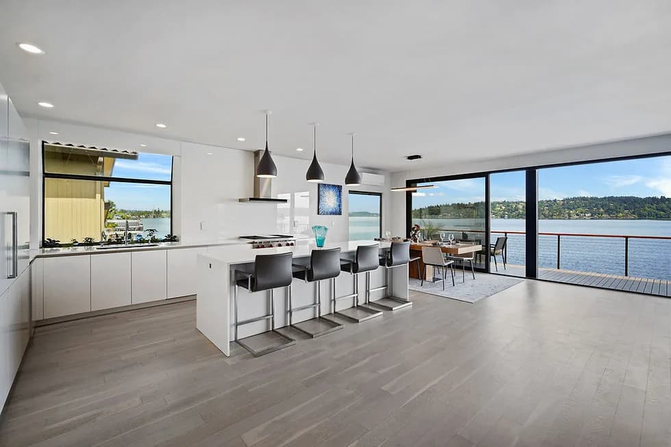 Modern open kitchen with island and bar stools, dining area, and large glass walls overlooking water
