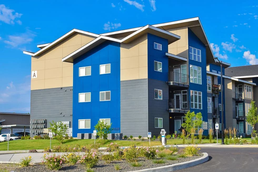 Three-story apartment building with blue central bays, beige and gray siding, balconies, and landscaped front