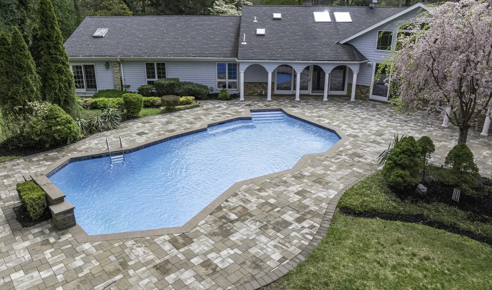Aerial view of a backyard pool surrounded by a stone patio and a covered arched porch along a grey house