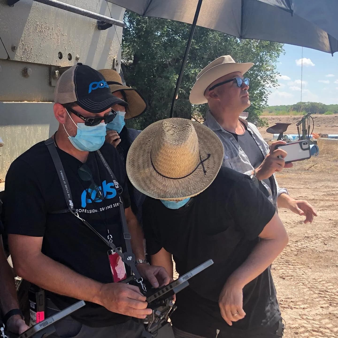 Three drone operators wearing hats and masks under an umbrella near a metal structure, with controllers