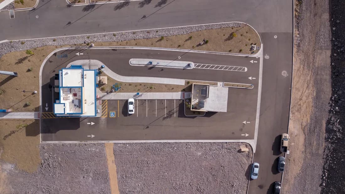 Top-down view of a small drive-through building, parking lot, and adjacent road with parked cars