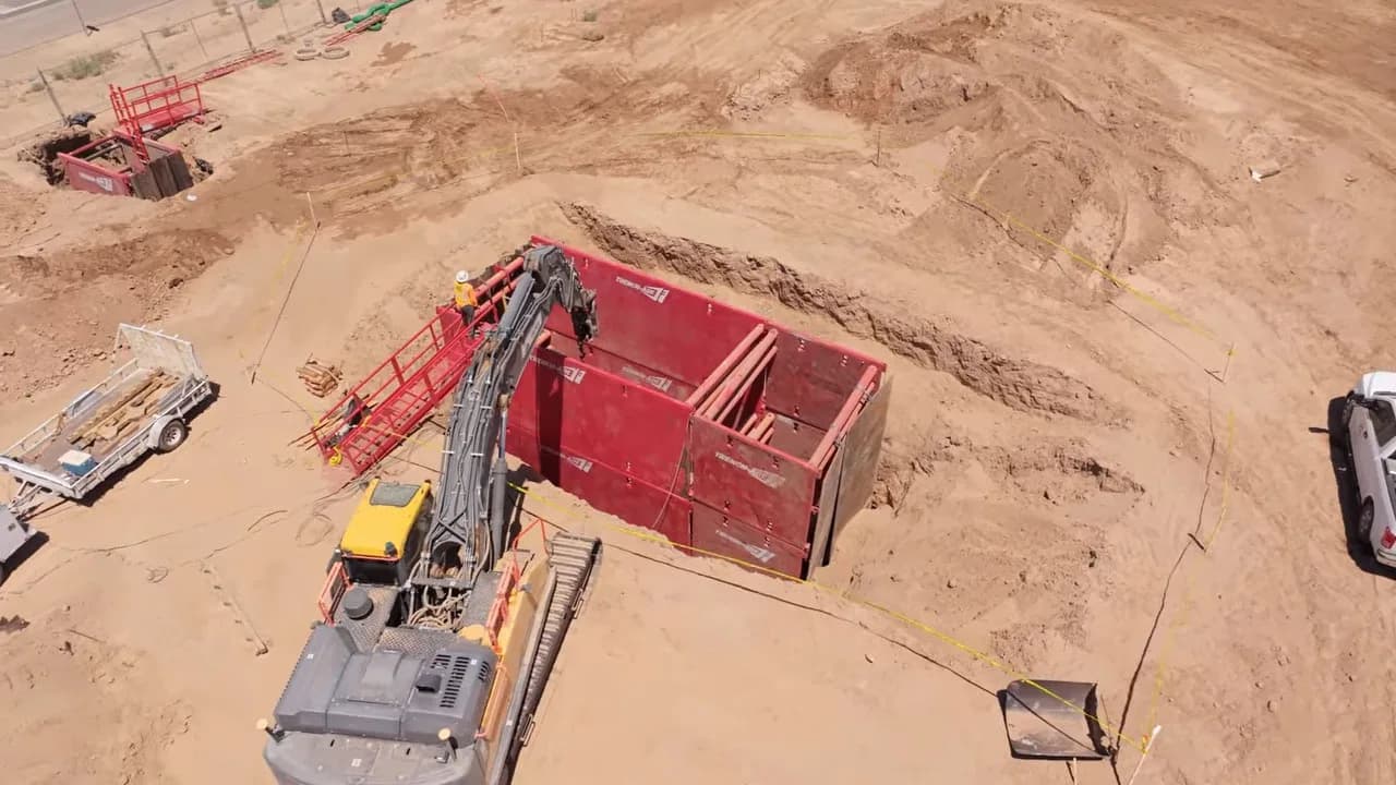 Aerial view of a construction excavation with excavator, red trench box and pickup truck