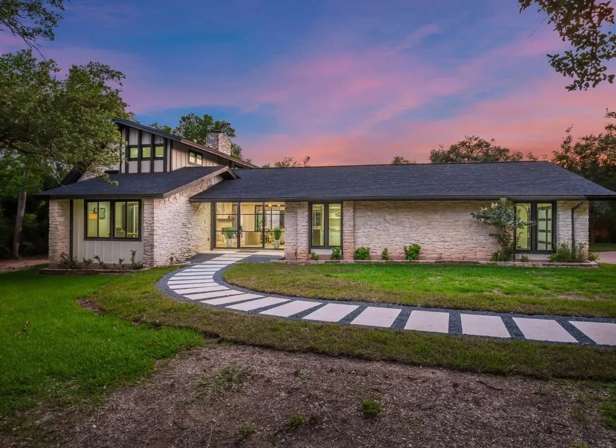 Modern stone house with large front windows and a curved stepping-stone path across a green lawn at dusk
