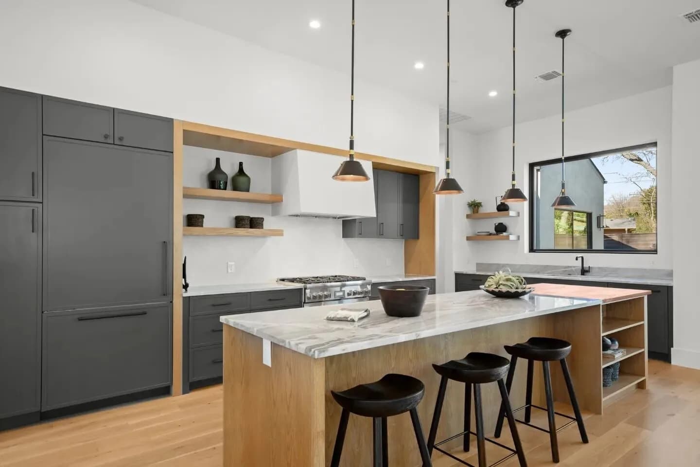 Modern kitchen with grey cabinets, marble island, bar stools, open shelves, and large window