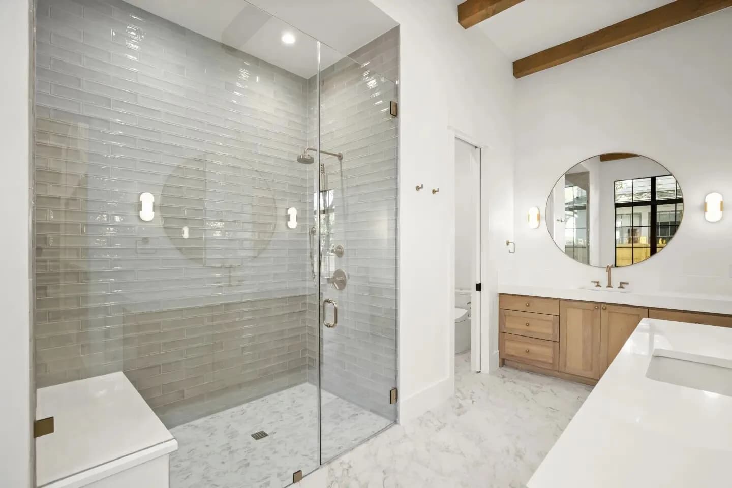 Glass shower enclosure with gray subway tiles, a bench, marble floor, and a wooden vanity with round mirror