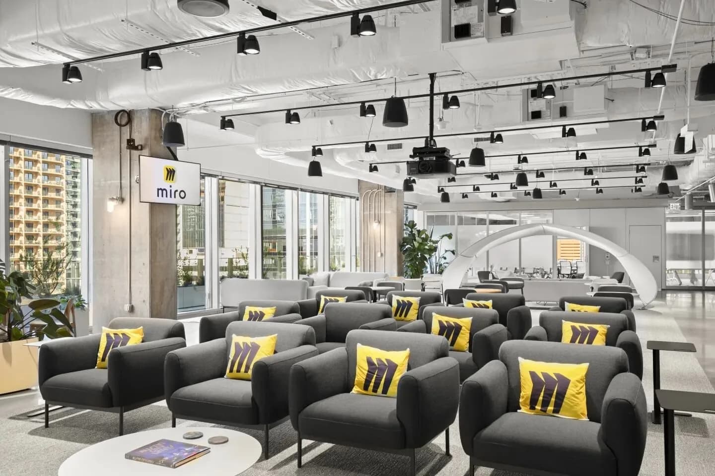 Modern lounge with gray armchairs and yellow pillows, Miro logo sign, curved white arch, projector, windows