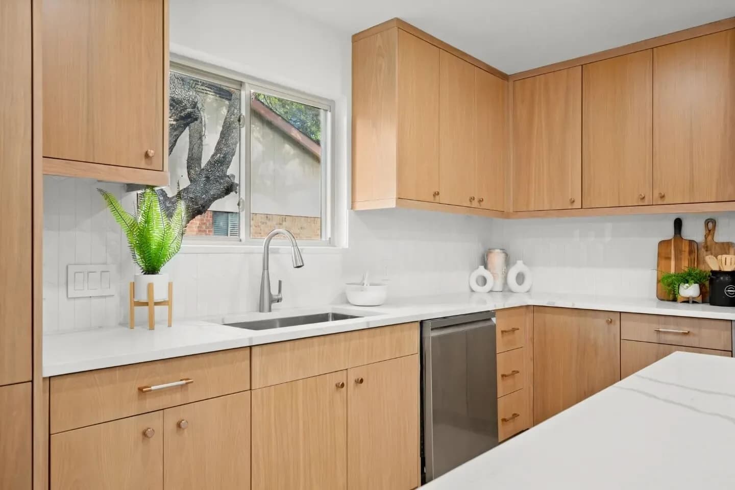 Kitchen with light wood cabinets, white countertops, sink and dishwasher, window with tree outside, fern