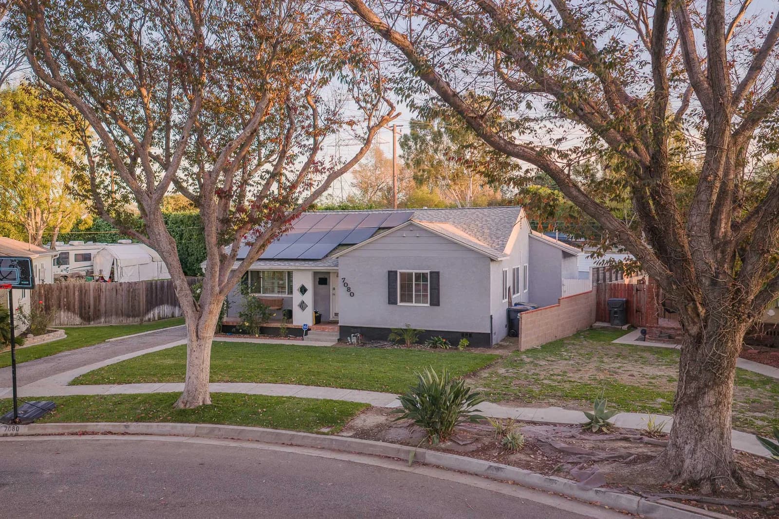 Residential street with a gray single-story house, solar panels, front yard, and large trees framing