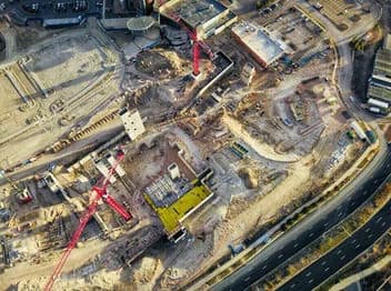 Aerial view of a construction site with red cranes, dirt piles, and surrounding roadways