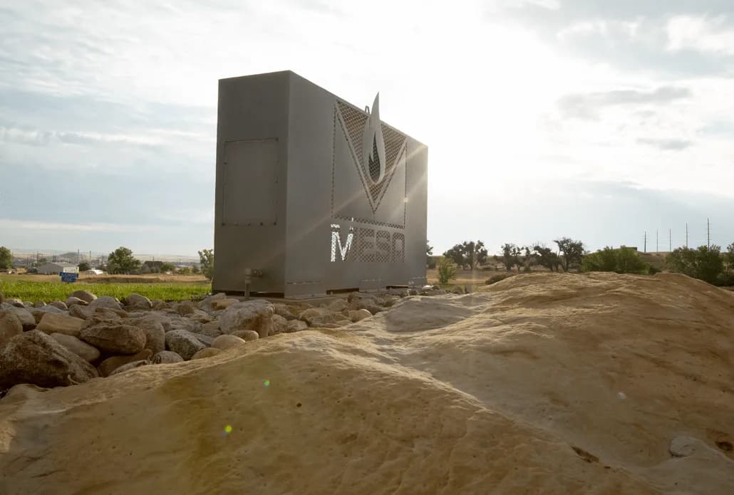 Large gray metal container with a logo on its side, placed on rocky ground with a sunny sky