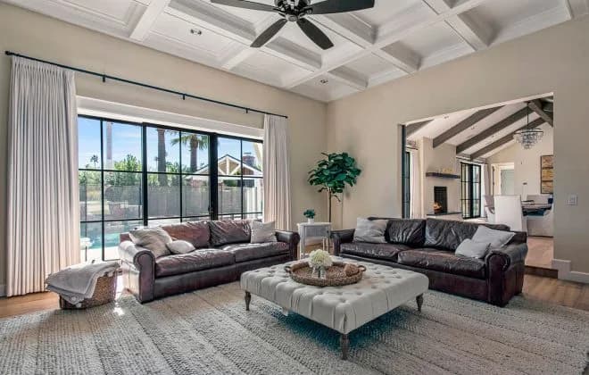 Bright living room with two leather sofas, tufted ottoman, coffered ceiling, and pool visible through large windows