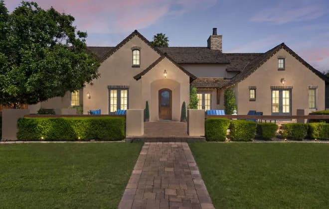 Evening frontal view of a stucco suburban house with lit windows, walkway, and manicured lawn